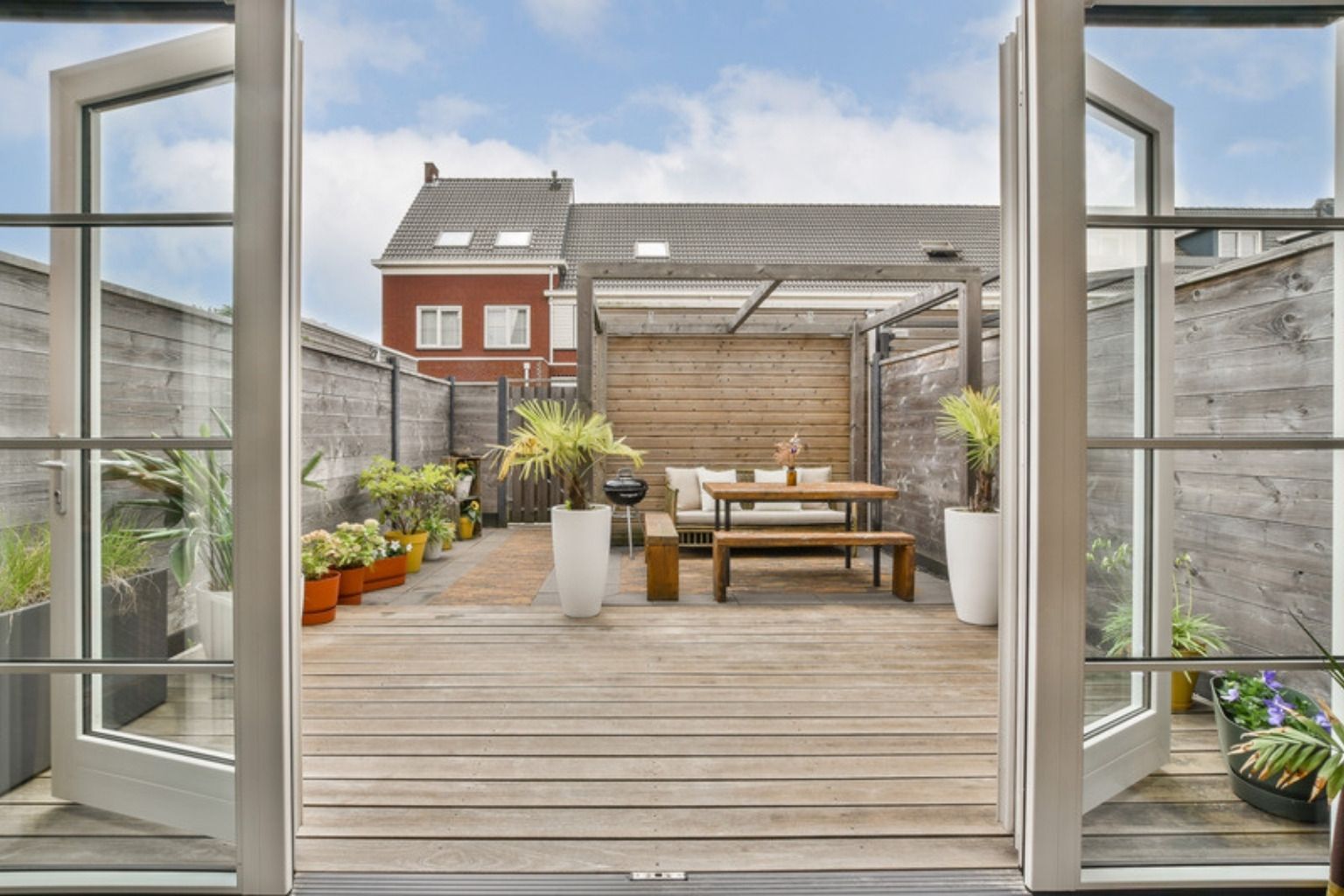 Træterrasse med spisebord og potteplanter set gennem åbne glasdøre, med røde murstenshuse i baggrunden