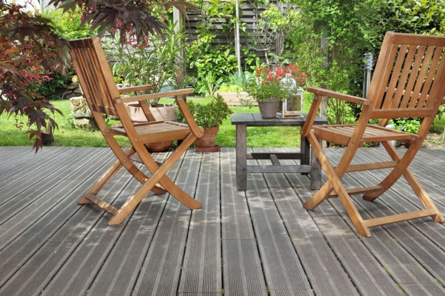 To træklapstole og et lille bord på en grålig træterrasse omgivet af planter og blomster