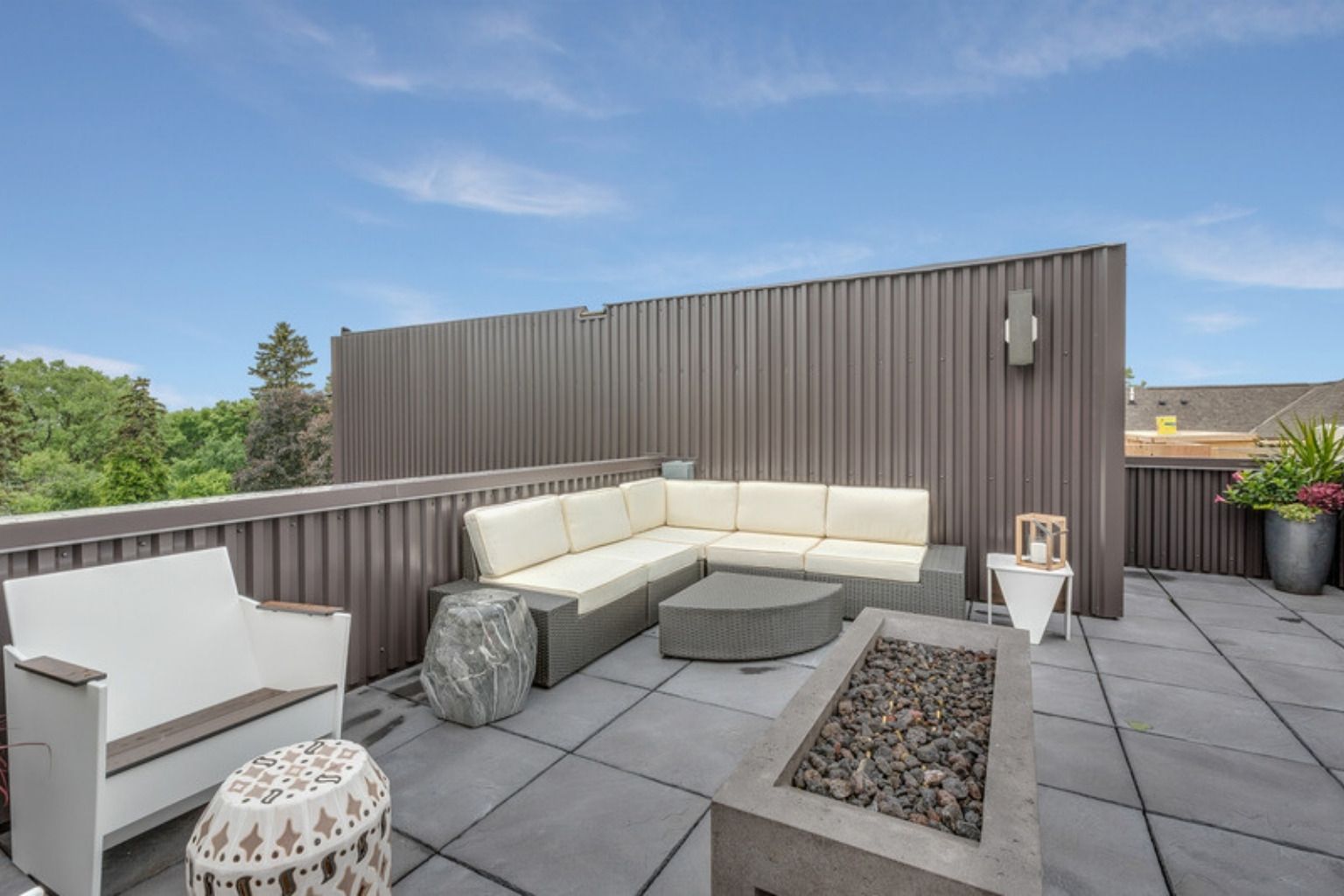 Tagterrasse med grå fliser, hvid loungesofa og bålbord, omgivet af trætoppe