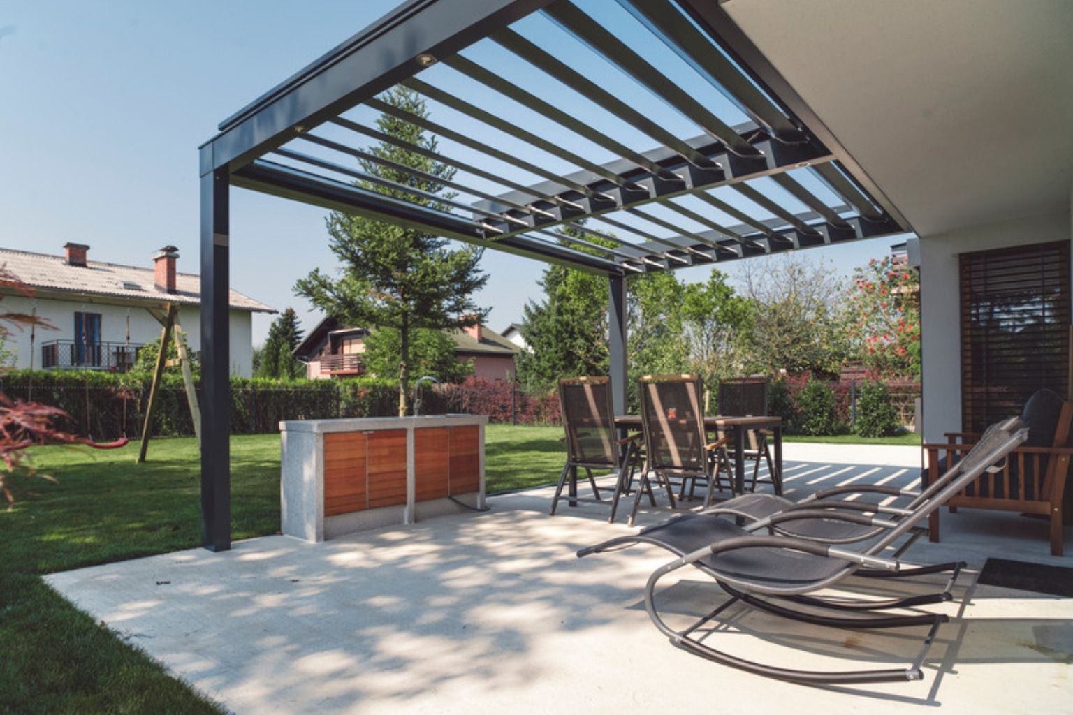 Sort lamelpergola i aluminium over en lys fliseterrasse med havemøbler og græsplæne